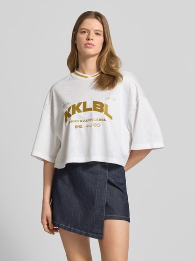 Frauenmodel trägt Cropped T-Shirt mit rückseitigem Logo-Print von Karo Kauer, weiß mit Label-Print, Loose-Fit, Rundhalsausschnitt, überschnittene Schultern, Rückenlänge 47 cm in Größe S, Ganzkörperansicht von vorne.