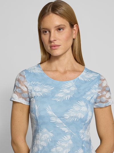 Kobiece model prezentuje Knielanges Kleid mit Rundhalsausschnitt od Apricot, mieszanka materiałów z dodatkiem streczu, taliowany krój, długość do kolan, okrągły dekolt, nieprześwitująca halka pod sukienkę, długość tyłu w rozmiarze S: 94 cm, szerokość w rozmiarze S: 46 cm, długość rękawów w rozmiarze S: 19 cm, niebieskie i białe wzory liści, faktura lekko przezroczysta na rękawach, ujęcie zbliżeniowe górnej części ciała.