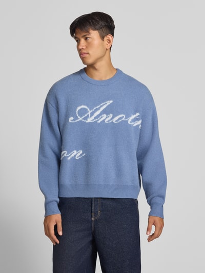 Męski model nosi sweter z okrągłym dekoltem 'Pullover mit geripptem Rundhalsausschnitt' od ANOTHER COTTON LAB, prosty krój, prążkowane wykończenia, długość tyłu w rozmiarze M: 61 cm, długość rękawów w rozmiarze M: 63 cm, odcienie niebieskiego, widoczne białe litery, gładka faktura, całkowity widok z przodu.