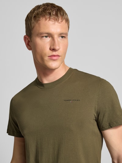 Männermodel trägt Slim Fit T-Shirt aus reiner Baumwolle von Tommy Jeans in Oliv Unifarben mit Label-Print, Nahaufnahme Oberkörper.