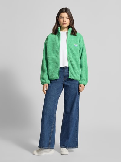 Frauenmodel trägt Jacke aus Teddyfell Modell 'HOKTOWN' von American Vintage in Grün mit Stehkragen, Reißverschluss, Label-Detail, Eingrifftaschen, Lockerer-Schnitt; Rückenlänge 65 cm, Schulterbreite 60 cm, Brustweite 59 cm, Ärmellänge 55 cm, Ganzkörperansicht von vorne.