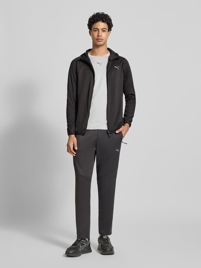 Männermodel trägt Regular Fit Trainingsjacke mit Logo-Print von PUMA PERFORMANCE in Schwarz mit Kapuze und Reißverschluss, Ganzkörperansicht von vorne.