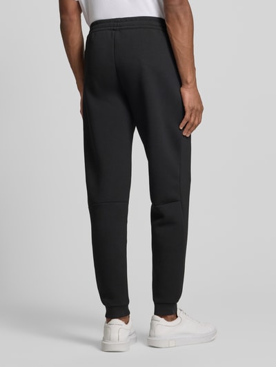 Männermodel trägt Sweatpants mit Viskose-Anteil Modell 'TRAIN PREMIUM SHIELD' von EA7 Emporio Armani in Schwarz, Gerade Passform, Gerippte-Abschlüsse, Elastischer Bund, Reißverschlusstaschen, Material-Mix, Ganzkörperansicht Rückansicht.