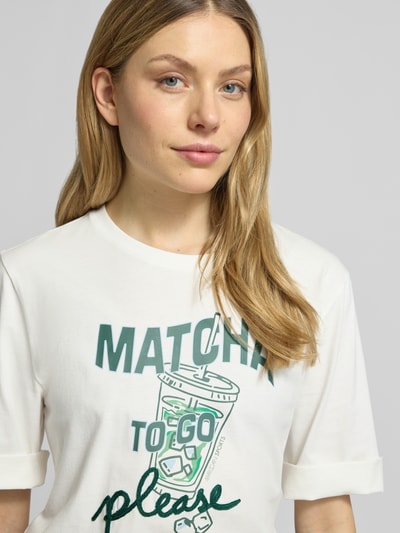 Frauenmodel trägt Slim Fit T-Shirt mit Motiv-Print von Marc Cain aus Reine Baumwolle, Logo-Print, Halbarm, Rundhalsausschnitt, Nahaufnahme Oberkörper.