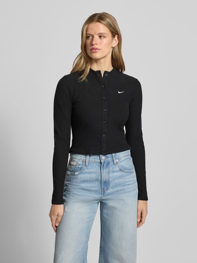 Frauenmodel trägt Cardigan in Ripp-Optik Modell 'CHILL' von Nike, Skinny-Fit, Stehkragen, Label-Stitching, Langärmelig, Durchgehende Knopfleiste, schwarze Farbe mit feiner Ripp-Struktur, Jeans kombiniert, Nahaufnahme Oberkörper