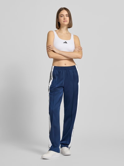 Frauenmodel trägt Straight Fit Jogginghose mit elastischem Bund Modell 'Adibreak' von adidas Originals in Blau mit weißen Streifen, Ziernähte und Eingrifftaschen, glatte Textur, Ganzkörperansicht von vorne.