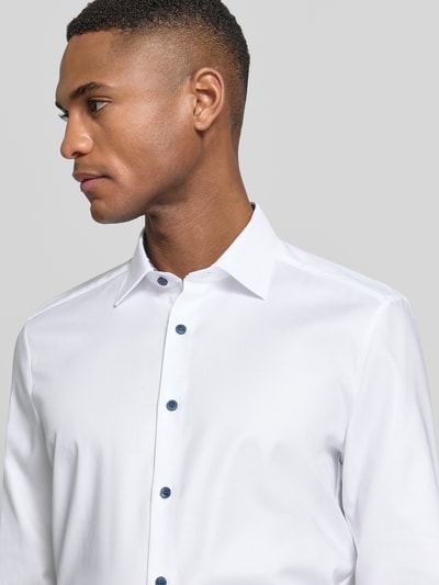 Męski model nosi Slim Fit Freizeithemd mit Kentkragen od OLYMP Level Five, biały, długi rękaw, niebieskie guziki, elastyczna bawełna, widok całej górnej części ciała z przodu. Szerokość w rozmiarze 40: 44 cm, długość tyłu: 80 cm, długość rękawa: 68 cm, krój slim fit, sportowe mankiety.