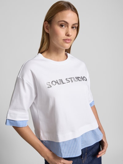 Kobiece model nosi T-Shirt mit Paillettenbesatz und Rundhalsausschnitt od Smith and Soul, damski t-shirt z bawełny, pudełkowy krój, okrągły dekolt, cekinowe obszycie, lekko opuszczone ramiona, długość tyłu w rozmiarze S: 54 cm, białe i niebieskie kolory, tekstura gladka, widok z przodu w półzbliżeniu.