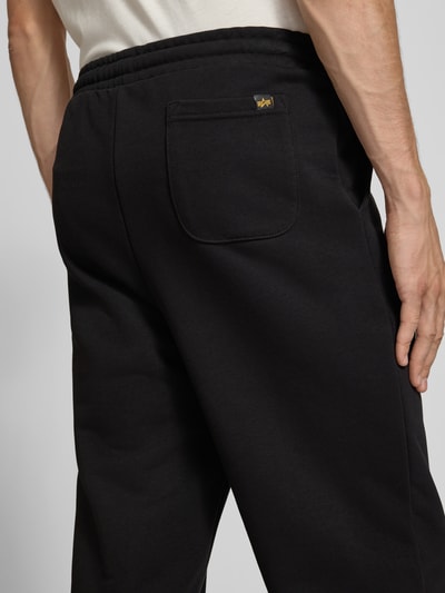 Männermodel trägt Sweatpants mit Label-Print von Alpha Industries in Schwarz, lockere Passform, Detailaufnahme Rückansicht, Baumwoll-Mix.