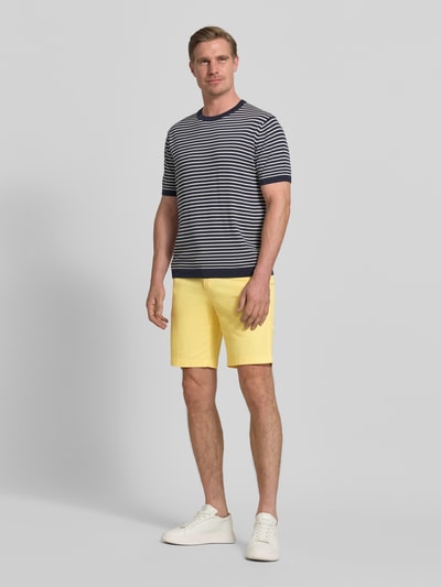 Męski model nosi Regular Fit Bermudas aus Baumwoll-Mix od Tommy Hilfiger, jednokolorowe, logo-stitching, boczne i tylne kieszenie, bawełniany materiał, żółty kolor, widok całej sylwetki z przodu.