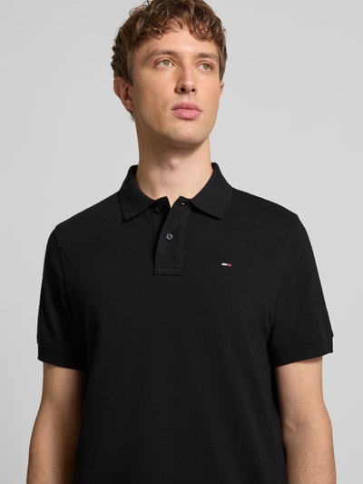 Męski model nosi Regular Fit Poloshirt aus reiner Baumwolle von Tommy Jeans, efekt: jednolity kolor, krój regular fit, wyhaftowane logo, kołnierzyk polo, krótki rękaw, wykończenie z rozcięciami po bokach, wykończenie materiału: pika, 100% bawełna, czarny kolor, półzbliżenie sylwetki z przodu.