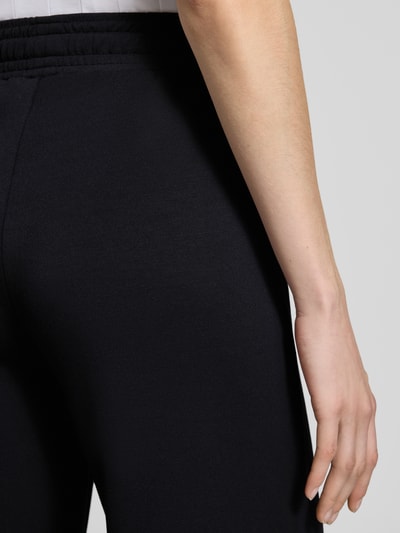 Kobiece modelka ma na sobie spodnie dresowe Wide Fit Sweatpants in verkürzter Passform Modell 'Banu' od Soyaconcept, elastyczna mieszanka materiałów z domieszką modalu, krój wide fit, skrócony, czarny kolor, detal – tunel w pasie, zbliżenie na biodra i dłoń, detal.