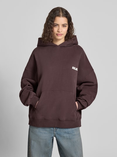 Frauenmodel trägt Oversized Hoodie mit Label-Prints und Känguru-Tasche von Karo Kauer, braune Baumwolle, lockere Form, Ganzkörperansicht von vorne.