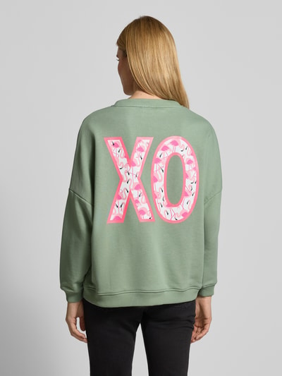 Frauenmodel trägt Relaxed Fit Sweatshirt mit Pailettenbesatz Modell 'XO' von miss goodlife, grün mit rosa Details, Rückansicht.