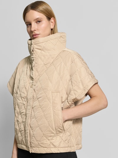 Frauenmodel trägt Cape mit Steppnähten und Eingrifftaschen von Copenhagen Muse in Beige, Relaxed-Fit, Knopfleiste, Hochgeschlossen, Nahaufnahme Oberkörper. Rückenlänge 66 cm, Schulterbreite 42 cm, Brustweite 25 cm bei Größe S/M.
