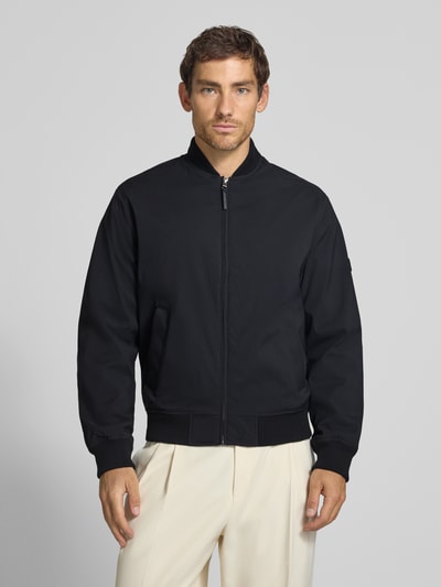Męski model nosi bomberkę z zamkiem błyskawicznym i wpuszczanymi kieszeniami Bomberjacke mit Reißverschluss und Eingrifftaschen von JACK & JONES, krój regular fit, stójka, długość tyłu w rozmiarze M: 66 cm, szerokość ramion w rozmiarze M: 42 cm, szerokość klatki piersiowej w rozmiarze M: 60 cm, długość rękawów w rozmiarze M: 68 cm, czarny kolor, gładka tekstura, całkowity widok z przodu.