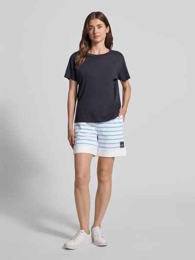Frauenmodel trägt Shorts mit Label-Detail und Tunnelzug von Marc Cain, weiß-blau gestreift, Baumwoll-Mix mit Modal-Anteil, Regular Fit, elastischer Bund, lockere Passform, Kombination mit dunkelblauem Shirt, Ganzkörperansicht von vorne.