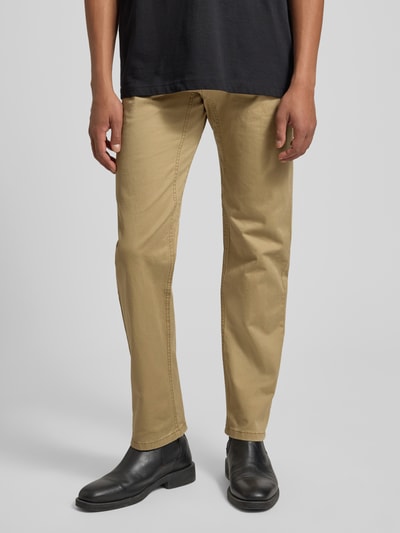 Mężczyzna nosi Regular Fit Chino aus Baumwoll-Mix von s.Oliver RED LABEL w jednolitym kolorze beżowym, bawełniany materiał z pięcioma kieszeniami i ukrytą kieszenią na zamek, guzik oraz szlufki na pasek, widok z przodu, ujęcie całej sylwetki.
