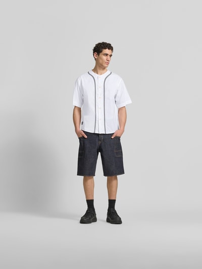 Männermodel trägt Freizeithemd in kurzärmeligem Design von Kenzo, Reine Baumwolle, Relaxed-Fit, Rundhalsausschnitt, Knopfleiste, weiße Grundfarbe mit feinen schwarzen Linien, kombiniert mit dunklen Jeansshorts, glatte Textur, Ganzkörperansicht von vorne.