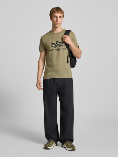 Männermodel trägt T-Shirt mit Logo und Rundhalsausschnitt von Alpha Industries in Olivgrün, Regular-Fit, Reine Baumwolle, Rückenlänge 68 cm, Ganzkörperansicht von vorne.