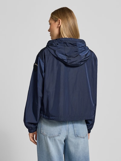 Frauenmodel trägt Trekking Jacke mit Kapuze Modell 'BURRAGE' von Blauer Usa in Dunkelblau, glatte Textur, Ganzkörperansicht Rückansicht. Elastische Ärmelabschlüsse, seitliche Reißverschlusstaschen, gerader Schnitt, Zweiwege-Reißverschluss, Label-Patch sichtbar.