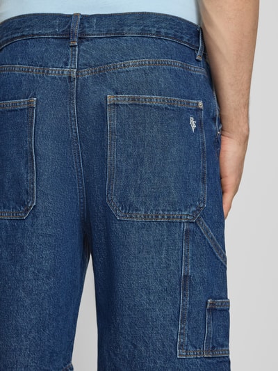 Männermodel trägt Baggy Fit Jeansshorts mit Eingrifftaschen von REVIEW aus Reine Baumwolle in dunkler Jeans-Optik, Detailaufnahme Rückansicht. Bundweite bei Größe M: 43 cm, Innenbeinlänge: 25 cm, Knopf- und Reißverschluss.