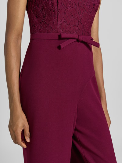 Frauenmodel trägt Regular Fit Jumpsuit mit Zierschleife von Adrianna Papell, florale Spitze, weinrote Farbe, Detailaufnahme, Material-Mix mit Viskose-Anteil.