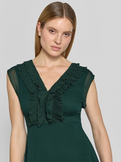 Modelka kobieca ma na sobie Knielanges Kleid mit Rüschen und V-Ausschnitt od Zero, szyfon, taliowany krój, długość do kolan, dekolt w serek, falbany, rękawy z falistym wykończeniem, zamek błyskawiczny z tyłu, długość tyłu w rozmiarze 36: 84 cm, szerokość w rozmiarze 36: 40 cm, głęboka zieleń, tekstura lekko prześwitująca, gładka tkanina. Zbliżenie górnej części ciała z przodu.