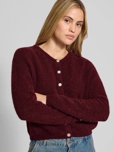 Kobiece model prezentuje kardigan Relaxed Fit Cardigan aus Woll-Alpaka-Mix Model 'VITOW' od American Vintage, mieszanka wełny i alpaki, imitacja dzianiny, listwa guzikowa, okrągły dekolt, kolor bordowy, długość tyłu 52 cm, długość rękawów 56 cm, ujęcie z przodu, zbliżenie na górną część ciała.
