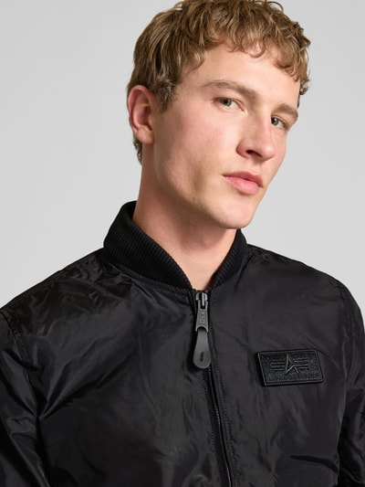 Männermodel trägt Blouson mit Logo-Anhänger Modell 'MA-1 TT' von Alpha Industries in Schwarz, Label-Patch, glatte Textur, Nahaufnahme Oberkörper. Herren, Lockere Passform, Reißverschluss, Armtasche, Elastische Abschlüsse:, Rückenlänge Größe M 69 cm, Schulterbreite 46 cm, Brustweite 54 cm, Ärmellänge 67 cm.