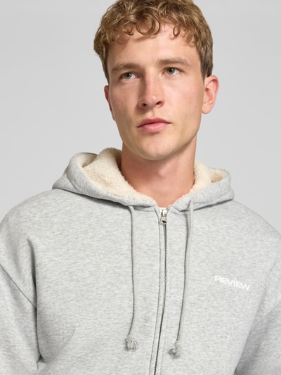 Männermodel trägt Regular Fit Sweatjacke aus Baumwoll-Mix mit Teddyfutter von REVIEW in Hellgrau, Logo-Detail, Nahaufnahme Oberkörper.