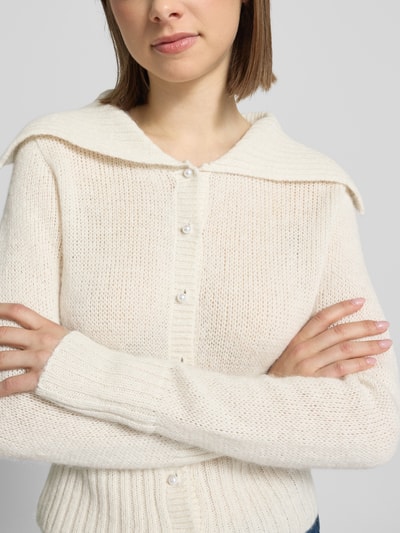 Kobiece model nosi kardigan rozpinany z szerokim kołnierzem i listwą guzikową od Gina Tricot, mieszanka materiałów z domieszką alpaki, krój regular fit, kremowy kolor, długość tyłu w rozmiarze S: 46 cm, długość rękawów w rozmiarze S: 60 cm, zbliżenie na górną część ciała.
