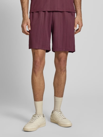 Männermodel trägt Shorts mit elastischem Bund Modell 'Insignia Shadow' von KARL KANI in Lila mit Streifenmuster, Relaxed-Fit, Ganzkörperansicht von vorne, Bundweite 37 cm, Innenbeinlänge 20 cm, Größe M.