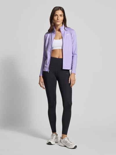 Frauenmodel trägt Motion Jacket mit Logo-Print Modell 'EMEA' von Under Armour in Lila, körpernaher Schnitt, Stehkragen, Reißverschluss, seitliche Reißverschlusstaschen, kombiniert mit Sportleggings und Sneakers, Ganzkörperansicht von vorne. Rückenlänge bei Größe S: 60 cm, Schulterbreite: 36 cm, Brustweite: 42 cm, Ärmellänge: 60 cm.