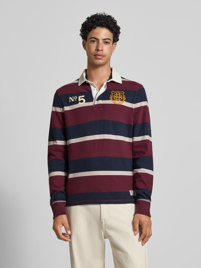 Männermodel trägt Regular Fit Rugby Shirt mit Logo- und Motiv-Stitching von Superdry in Dunkelblau, Rot und Creme, Reine Baumwolle, Polokragen, Knopfleiste, Label-Patch, Rückenlänge bei Größe M 70 cm, Ganzkörperansicht von vorne.