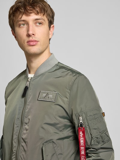 Männermodel trägt Regular Fit Bomberjacke mit Logo-Patch von Alpha Industries in Olivgrün, sichtbarer Reißverschluss, Nahaufnahme Oberkörper. Rückenlänge 65 cm, Schulterbreite 45 cm, Brustweite 54 cm, Ärmellänge 69 cm.