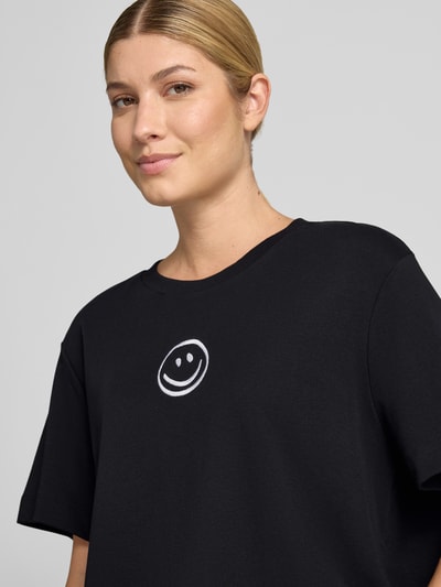 Kobiece modelka nosi czarny damski t-shirt z białym nadrukiem uśmiechu, gładka faktura, model 'SARA' od VERO MODA, zbliżenie na górną część ciała.