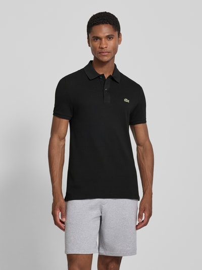 Męski model nosi czarną koszulkę polo Slim Fit Poloshirt aus reiner Baumwolle von Lacoste, prosty krój, gładka struktura, Ganzkörperansicht von vorne.