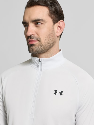 Mężczyznamodel nosi Sweatshirt mit kurzem Reißverschluss und Logo-Print von Under Armour w kolorze jasnoszarym z czarnym nadrukiem z logo, prosty krój, stójka, zamek błyskawiczny, długość tyłu w rozmiarze M: 74 cm, długość rękawów w rozmiarze M: 64 cm, zbliżenie na górną część tułowia.