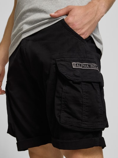 Męski model nosi szorty z kieszeniami cargo model 'CREW SHORT' od Alpha Industries, mieszanka bawełny i elastanu, krój regular fit, czarny kolor, zbliżenie na górną część nogawki.