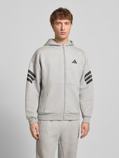 Männermodel trägt Sweatjacke mit Kapuze und Logo von adidas Sportswear, grau, Material-Mix mit Baumwoll-Anteil, Relaxed-Fit, Känguru-Tasche, Reißverschluss, Logo-Print, Ganzkörperansicht von vorne.