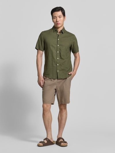 Mannelijk model draagt Slim Fit Freizeithemd aus Leinen-Baumwoll-Mix Modell 'AKSEL' van Casual Friday in olijfgroen, kentkraag, knoopsluiting, labelstiksel, korte mouwen, gecombineerd met beige short, volledige lichaamsaanzicht van voren, context: heren vrijetijdshemd casual friday, linnen-katoen mix, slim fit, kentkraag, label-stitching, knoopsluiting, ruglengte maat M 78 cm, schouderbreedte 45 cm, mouwlengte 27 cm.