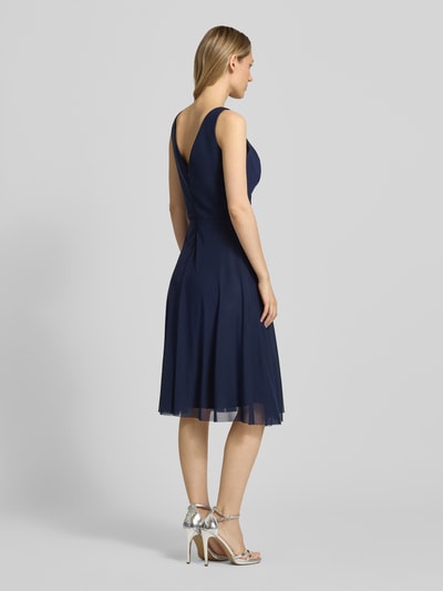 Frauenmodel trägt Cocktailkleid in Wickel-Optik mit V-Ausschnitt von TROYDEN COLLECTION, Mesh, taillierter Schnitt, Midikleid, Drapierungen, Rückenlänge bei Größe 36: 108 cm, Brustweite bei Größe 36: 41 cm, dunkles Blau, weich fließende Textur, Ganzkörperansicht Rückansicht.