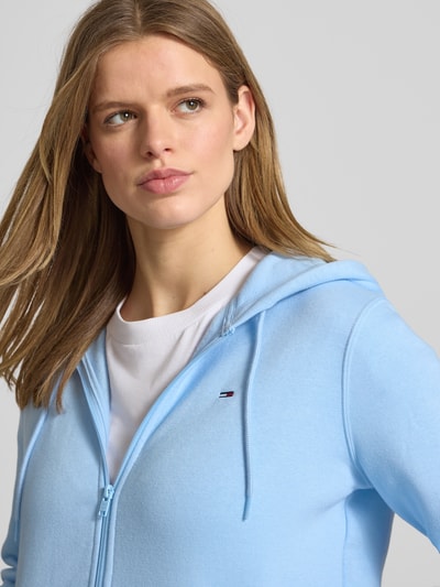 Frauenmodel trägt Regular Fit Sweatjacke aus Baumwoll-Mix von Tommy Jeans in Hellblau, unifarben, Kapuze, Logo-Stitching, Nahaufnahme Oberkörper.