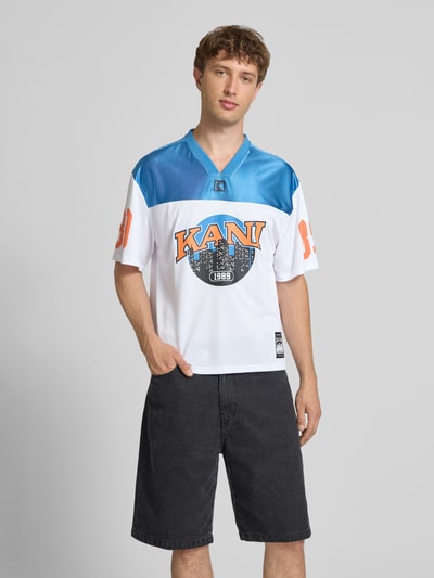 Männermodel trägt Jersey-Shirt mit V-Ausschnitt, Logo- und Motiv-Prints, Boxy-Fit von KARL KANI, Rückenlänge 64 cm, Ganzkörperansicht von vorne. Modell 'OG Sport College', Herren T-Shirt, College-Look, Label-Patch.
