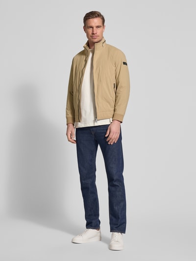 Männermodel trägt Regular Fit Jacke mit Opstaande kraag van bugatti in beige met tweewegritssluiting, steekzakken, logodetail, elastische mouwboord, capuchon, ruglengte 65 cm, schouderbreedte 47 cm, borstwijdte 53 cm, mouwlengte 63 cm; ganzkörperansicht von voren.