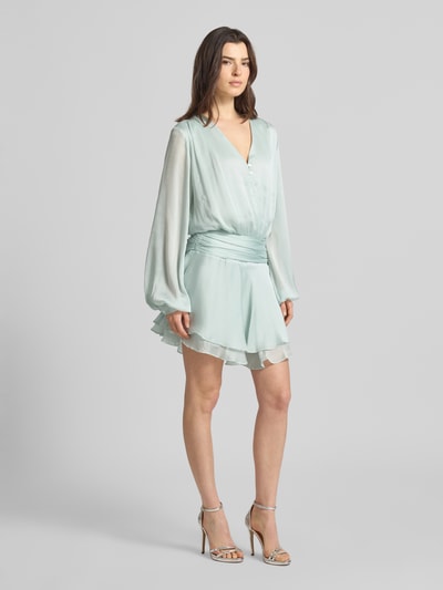 Frauenmodel trägt Minikleid mit kurzer Knopfleiste Modell 'MAGALI' von Guess in Mintgrün, Volantsaum, Taillierter Schnitt, V-Ausschnitt, seitlicher Knopf- und Reißverschluss, Ganzkörperansicht von vorne.