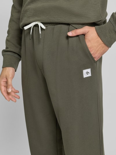Männermodel trägt Sweatpants mit elastischem Bund und Tunnelzug von Scotch & Soda in Olivgrün, Logo, Nahaufnahme Oberkörper, Regular Fit, reine Baumwolle, Innenbeinlänge Größe M 78 cm.