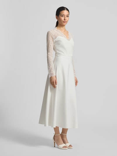 Frauenmodel trägt Tailliertes Brautkleid mit V-Ausschnitt Modell 'Joanna' von GUIDO MARIA KRETSCHMER WOMAN, Elastischer-Material-Mix, Oberteil aus Spitze, langärmelig, blickdichter Rockteil, Produkt in Creme, Ganzkörperansicht von vorne.