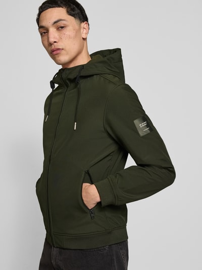 Mężczyznamodel nosi kurtkę softshell 'Softshelljacke mit Reißverschlusstaschen' od Jack & Jones w kolorze ciemnozielonym, krój regular fit, kaptur z tunelem, zamek błyskawiczny, kieszenie zapinane na zamek błyskawiczny, długość tyłu w rozmiarze M: 59 cm, szerokość ramion w rozmiarze M: 39 cm, szerokość klatki piersiowej w rozmiarze M: 48 cm, długość rękawów w rozmiarze M: 62 cm, gładka tekstura. Widok z przodu w ujęciu do pasa.